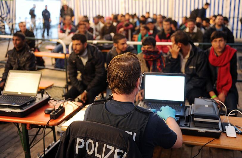 Ein Polizist sitzt am Passauer Bahnhof vor einem Computer. Im Hintergrund warten Flüchtlinge auf die Erfassung ihres Fingerabdrucks.