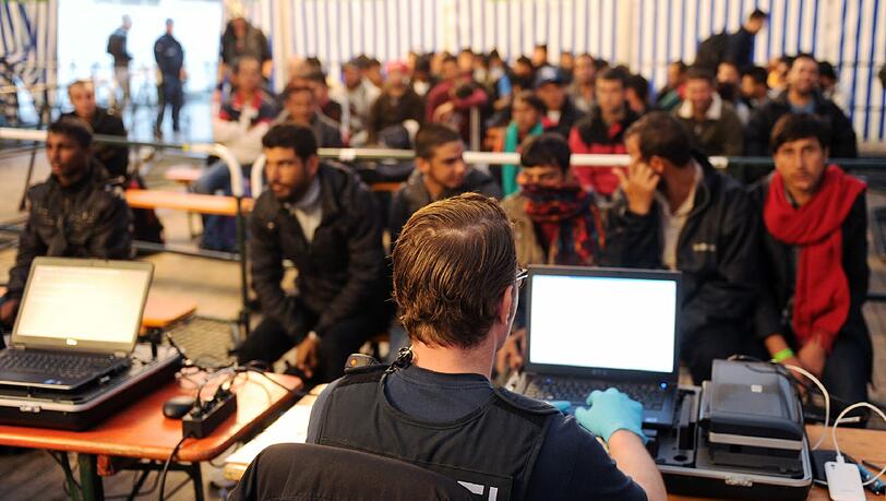 Ein Polizist sitzt am Passauer Bahnhof vor einem Computer. Im Hintergrund warten Flüchtlinge auf die Erfassung ihres Fingerabdrucks.