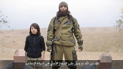 An der Seite eines schwer bewaffneten ISIS-Kämpfers tötet der 14-Jährige die beiden Männer, während sie im sandigen Boden knien.