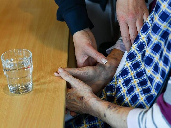 Eine Pflegerin hilft einer alten Frau, die an einem Tisch vor einem Glas Wasser sitzt. Foto: Jens Kalaene/zb/dpa
