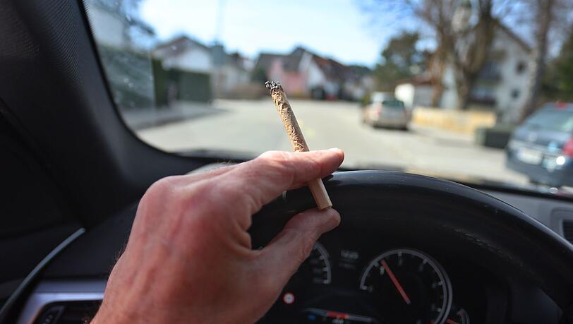 Ein Mann sitzt mit einem Joint zwischen den Fingern am Steuer eines Autos.