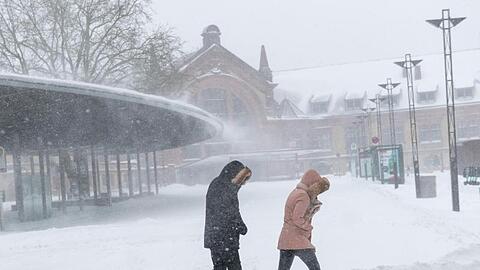 Zwei Menschen gehen mit eingezogenen Köpfen durch starken Schneefall mit Windböen vor dem Osnabrücker Hauptbahnhof.