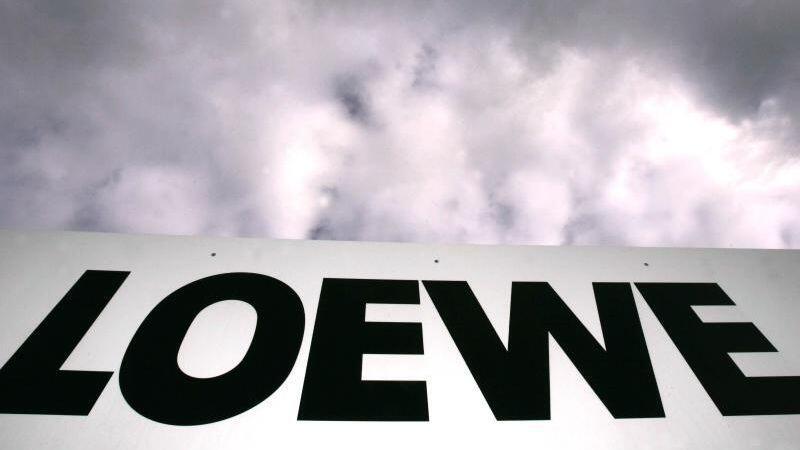 Das Loewe-Firmenlogo hängt vor dem bewölkten Himmel am Zaun des Betriebsgeländes in Kronach.