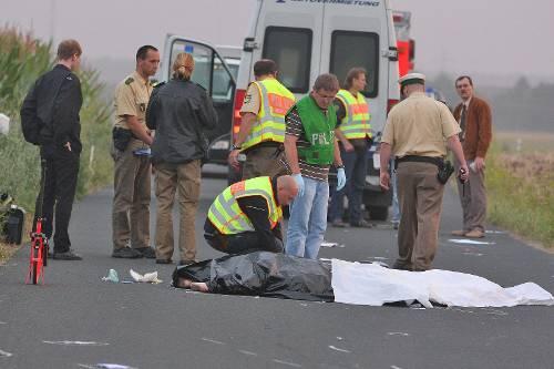 Die Leichen liegen abgedeckt auf der Straße. Die Polizei sichert die Spuren. Hinten steht der Unfallwagen.
