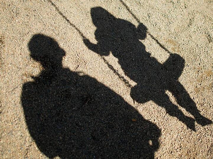 Der Schatten von einem Mann und einem schaukelnden Kind fallen auf Sand auf einem Spielplatz.