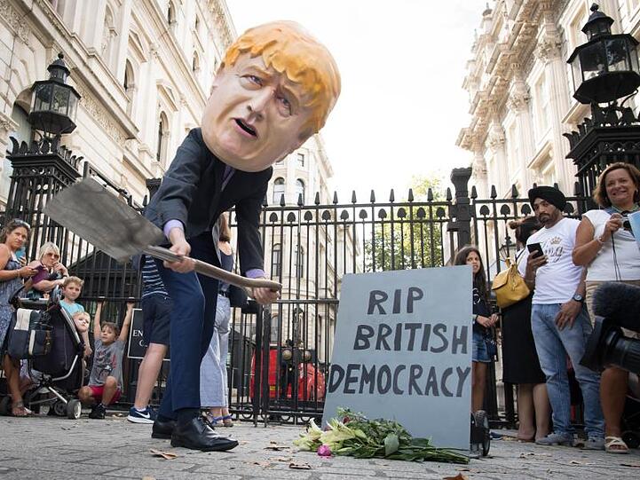 Ein Mann verkleidet mit einem riesigen Kopf des britischen Premierministers Johnson neben einem symbolischen Grabstein mit der Aufschrift "RIP British Democracy" (Ruhe in Frieden britische Demokratie).