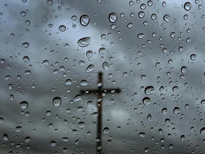 Ein Kreuz steht vor bedecktem Himmel, fotografiert durch eine regennasse Fensterscheibe. Foto: Karl-Josef Hildenbrand/Archivbild