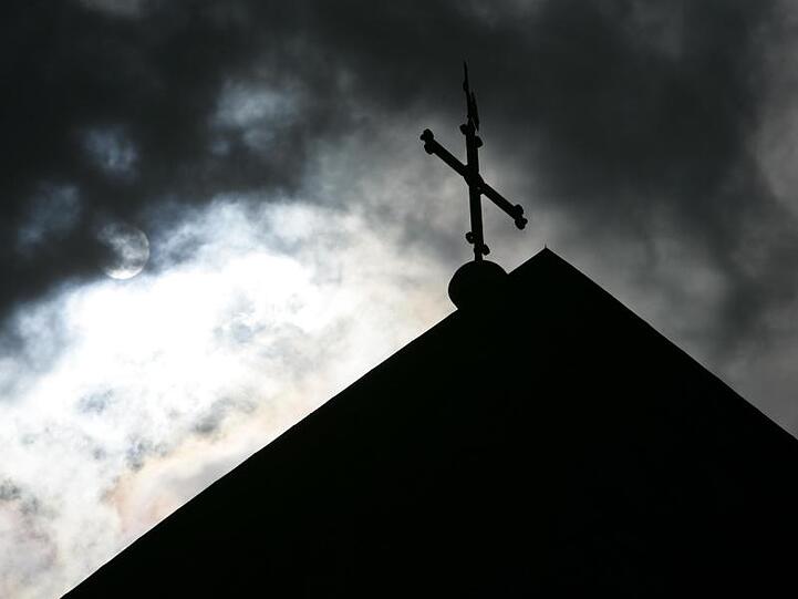 Im Gegenlicht und vor wolkenverhangenem Himmel ist die Kirchturmspitze eines Doms mit Kreuz zu sehen. Foto: Friso Gentsch/Archivbild