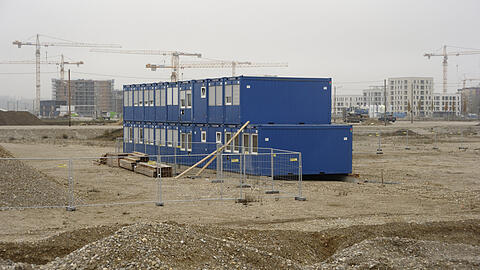 Winter 2020/21: Blaue Container, die für Bauarbeiter aufgestellt sind, stehen im Kies, der Humus ist abgeräumt. Monatelang geht hier nichts voran. Im Hintergrund aber wachsen die ersten Wohnhäuser.