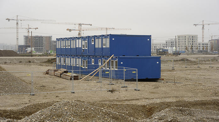 Winter 2020/21: Blaue Container, die für Bauarbeiter aufgestellt sind, stehen im Kies, der Humus ist abgeräumt. Monatelang geht hier nichts voran. Im Hintergrund aber wachsen die ersten Wohnhäuser.