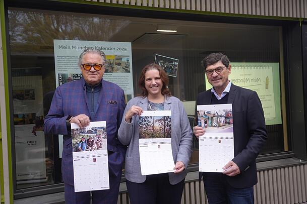 Schauspieler Michael Brandner (l.), Kommunalreferentin Jacqueline Charlier und der Projektinitiator Matthias Müller präsentieren den Fotokalender vor dem Standl 19 am neuen Elisabethmarkt. Das gemeinnützige Projekt "Mein München 2025" darf das städtische Standl, in das erst im Januar ein Marktgeschäft einzieht, bis 30. Dezember als Zwischennutzung nutzen.