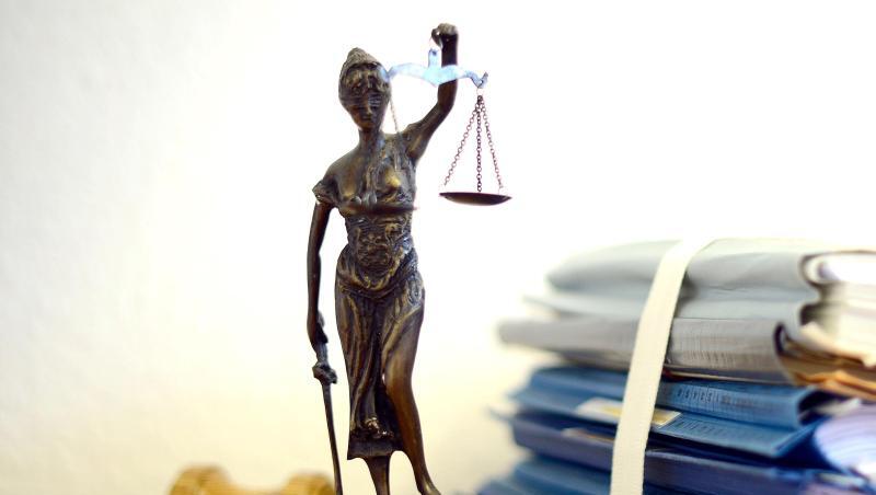 Eine Statue der Justitia steht neben Aktenbergen.