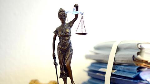 Eine Statue der Justitia steht neben Aktenbergen.