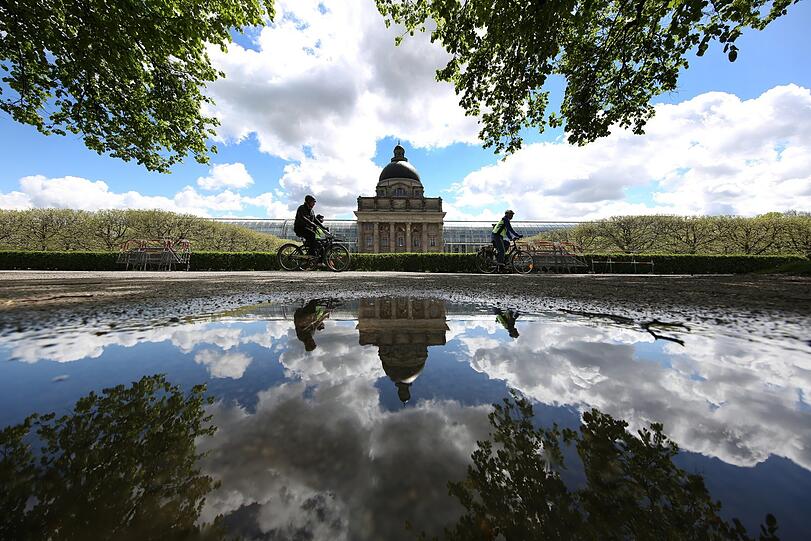 Was für ein Kunstwerk im Hofgarten: Staatskanzlei, Radler, Wolken und blauer Himmel spiegeln sich in einer Pfütze.