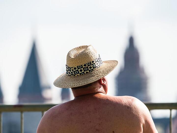 Ein Mann sitzt bei am Rheinufer mit Blick auf den Mainzer Dom und schützt sich mit einem Hut gegen die Sonne.