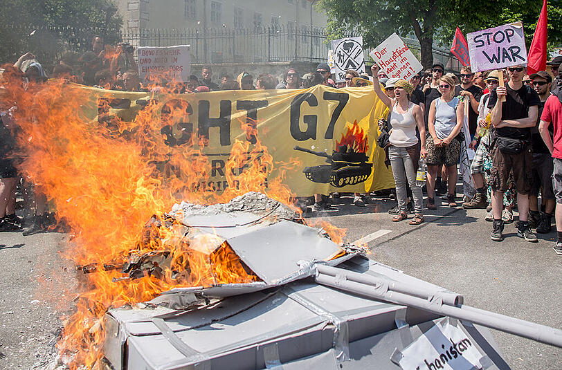 Fotografen, Reporter und Kameraleute umringen einen brennenden Panzer aus Pappe, auf dem die Ländernamen der Krisenherde Lybien, Mali, Kurdistan und Ukraine stehen, während einer Demonstration vor dem Marshall Center for European Security Studies in Garmisch-Partenkirchen.
