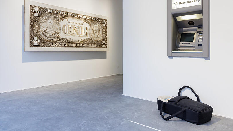 Eine Tragetasche samt Baby steht vor dem Geldautomaten. Hat da jemand sein Kind vergessen, oder spekuliert er oder sie, dass sich ein freundlicher Mitmensch erbarmt und dem &bdquo;Modern Moses&ldquo; ein gutes neues Zuhause schenkt? Was z&auml;hlt, ist das Geld. Tom Sachs bringt das mit seiner &uuml;berdimensionalen &bdquo;One Dollar&ldquo;-Note prima zum Ausdruck.