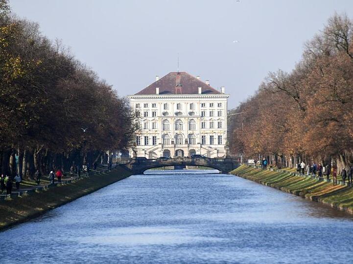 Spaziergänger spazieren auf den Wegen am Nymphenburger Kanal entlang. Im Hintergrund steht das Schloss Nymphenburg. Foto: Tobias Hase/Archiv