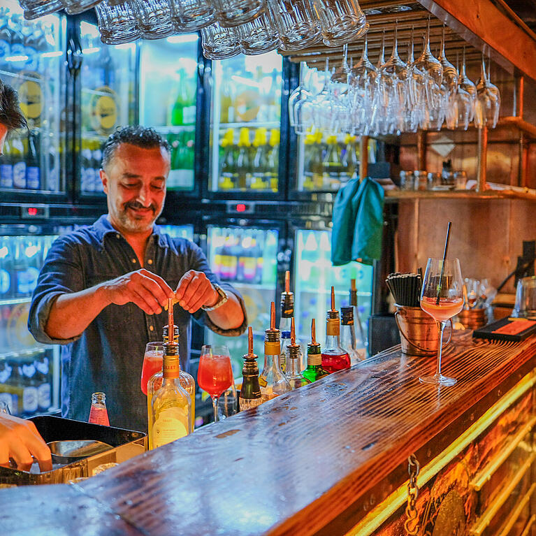 Vor drei Jahren er&ouml;ffnete Yilmaz Demir (Mitte) die Boazn in einem ehemaligen Kloh&auml;uschen an der Ludwigsbr&uuml;cke. Mittlerweile ist die Bar, die zugleich Tanzbar ist, vom Isarufer kaum mehr wegzudenken. Gerade mixt Demir die neue Spritzvariante "GrapefruitSpritz" f&uuml;r seine Stammg&auml;stin Katinka Behnke (rechts).