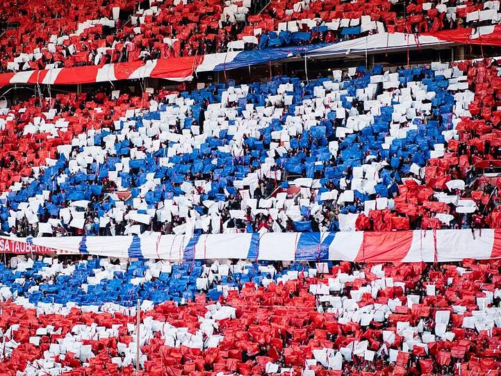 Fans halten blaue, weiße und rote Zettel hoch, die zusammen das Logo des FC Bayern ergeben. Foto: Matthias Balk/dpa/Archivbild