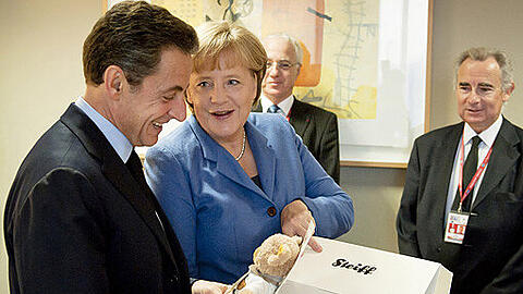 Baby-Geschenk: Angela Merkel überreicht Sarkozy hinter den Kulissen einen Teddybären für die kleine Giulia.