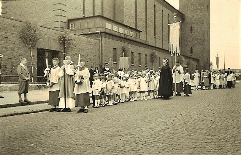 1938: Kindergartenkinder bei der Prozession vor der Kirche St. Sebastian in der noch kopfsteingepflasterten Karl-Theodor-Straße: Auch die Mutter von AZ-Leserin Gertraud Remmers ist zu sehen: das kleine Mädchen unterhalb der vordersten Fahne, das etwas abgelenkt von der Sache zur Seite schaut.