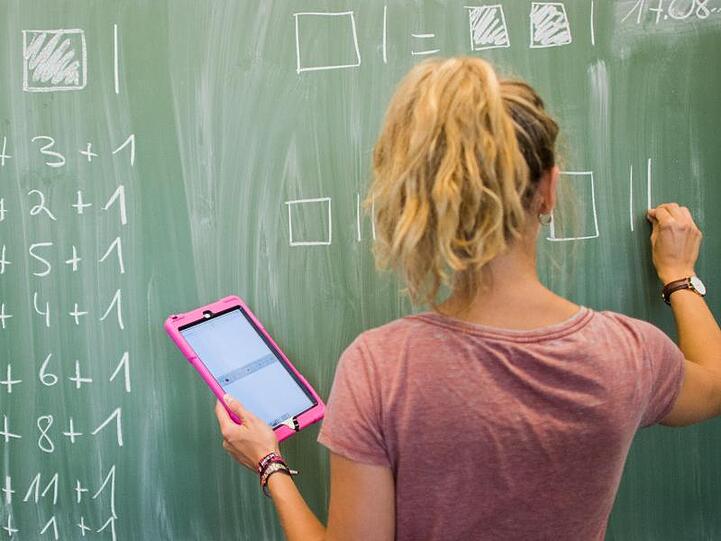 Eine Lehrerin schreibt an eine Schultafel und blickt dabei auf einen Tablet-Computer iPad. Foto: Julian Stratenschulte/Archiv