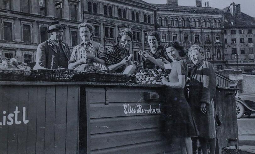 Um 1950 haben die Blumen- und Schwammerl-Marktfrauen am Viktualienmarkt noch grüne Kisten, an denen sie verkaufen. Links ist das kriegszerstörte Kustermann-Haus zu sehen, rechts ein Auto - die Straße führt zwischen den Ständen durch.