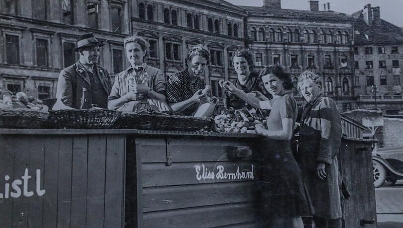 Um 1950 haben die Blumen- und Schwammerl-Marktfrauen am Viktualienmarkt noch grüne Kisten, an denen sie verkaufen. Links ist das kriegszerstörte Kustermann-Haus zu sehen, rechts ein Auto - die Straße führt zwischen den Ständen durch. Um 1950 haben die Blumen- und Schwammerl-Marktfrauen am Viktualienmarkt noch grüne Kisten, an denen sie verkaufen. Links ist das kriegszerstörte Kustermann-Haus zu sehen, rechts ein Auto - die Straße führt zwischen den Ständen durch.