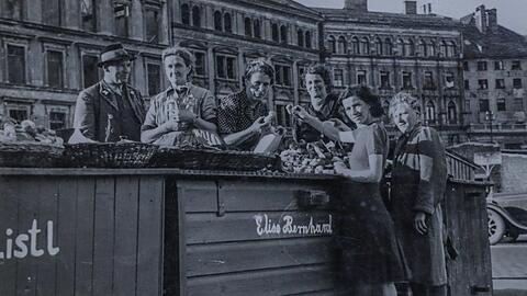 Um 1950 haben die Blumen- und Schwammerl-Marktfrauen am Viktualienmarkt noch grüne Kisten, an denen sie verkaufen. Links ist das kriegszerstörte Kustermann-Haus zu sehen, rechts ein Auto - die Straße führt zwischen den Ständen durch.