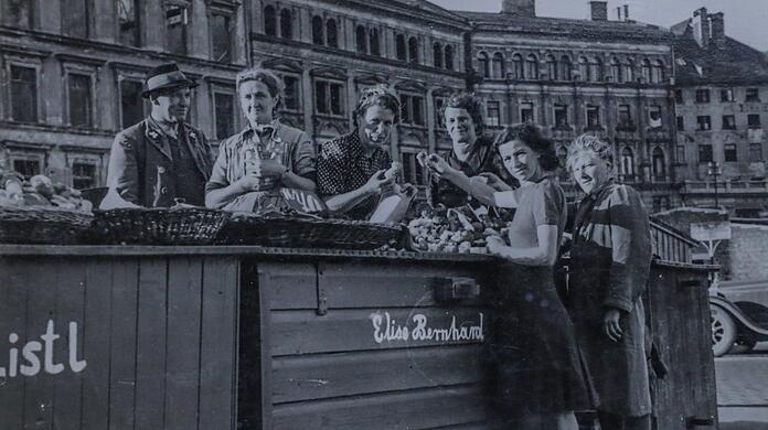 Um 1950 haben die Blumen- und Schwammerl-Marktfrauen am Viktualienmarkt noch grüne Kisten, an denen sie verkaufen. Links ist das kriegszerstörte Kustermann-Haus zu sehen, rechts ein Auto - die Straße führt zwischen den Ständen durch.
