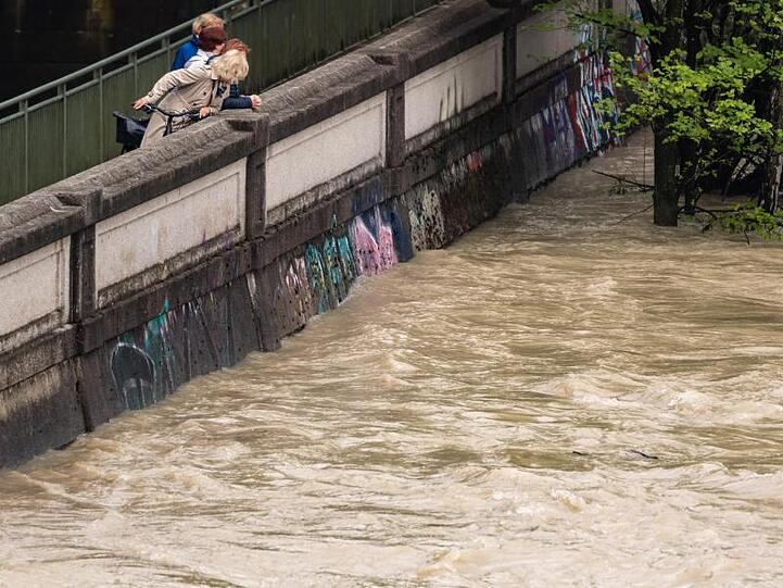 Passanten schauen von einem Gehweg zwischen dem Auer Mühlbach und der hochwasserführenden Isar auf das braune Wasser. Foto: Peter Kneffel