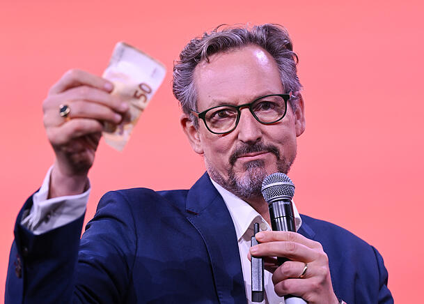 Eckart von Hirschhausen, Arzt und Wissenschaftsjournalist, hebt auf der Frankfurter Buchmesse einen Geldschein hoch. Auch auf der Pressekonferenz zu seiner neuen WDR-Doku nutzt er dieses Beispiel, um zu zeigen, wie die Deepfake-Betr&uuml;ger Vertrauen in gefestigte Institutionen missbrauchen.