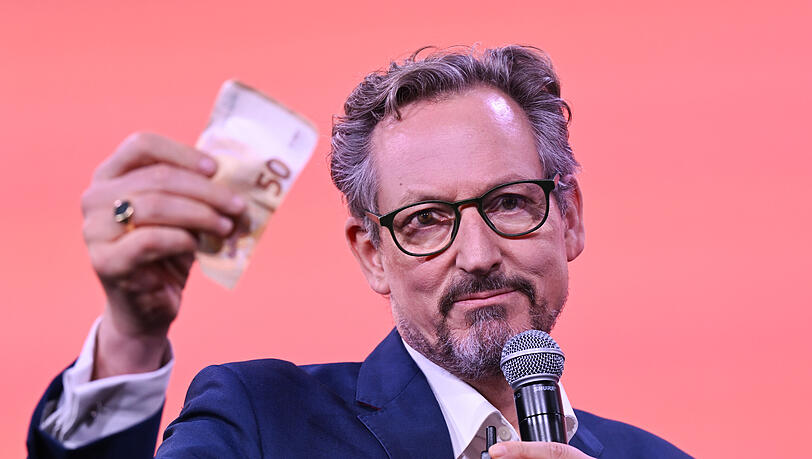 Eckart von Hirschhausen, Arzt und Wissenschaftsjournalist, hebt auf der Frankfurter Buchmesse einen Geldschein hoch. Auch auf der Pressekonferenz zu seiner neuen WDR-Doku nutzt er dieses Beispiel, um zu zeigen, wie die Deepfake-Betr&uuml;ger Vertrauen in gefestigte Institutionen missbrauchen.