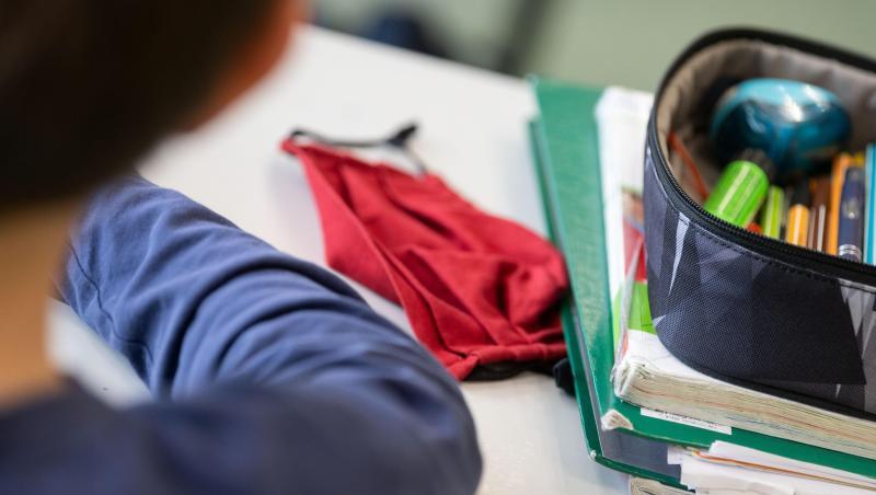 Eine Mund-Nasen-Bedeckung liegt während einer Unterrichtsstunde neben einem Mäppchen und Schulbüchern auf einem Schultisch.