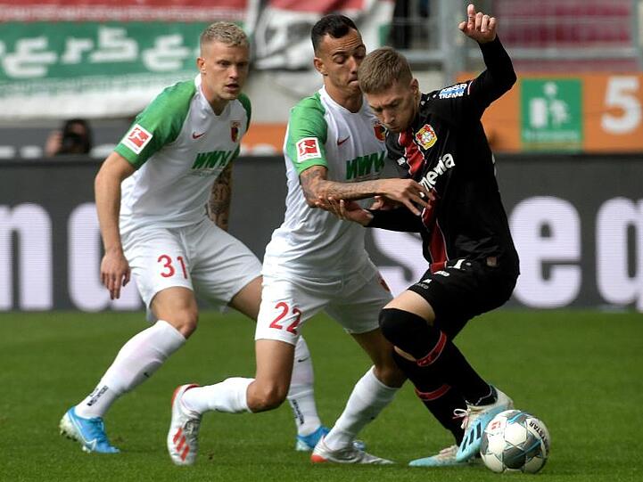 Augsburgs Iago (M) , Philipp Max (l) und Leverkusens Mitchell Weiser (r) kämpfen um den Ball.