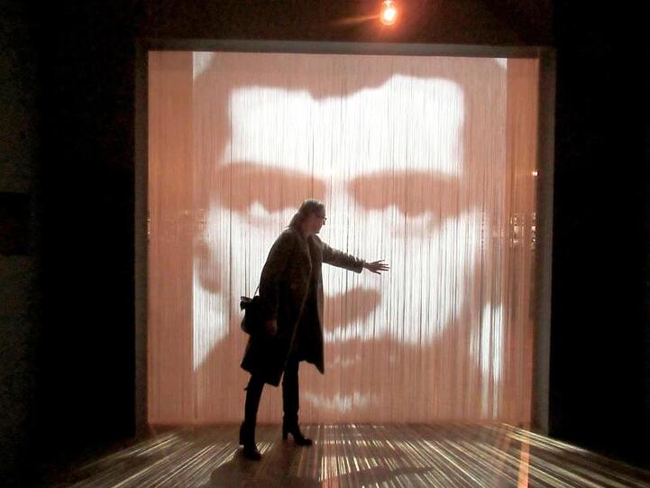Eine Besucherin steht vor einem Kunstwerk, einem Streifenvorhang mit einem Porträt des französischen Konzeptkünstler Christian Boltanski.
