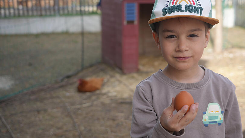 Mathis (6) hat gerade ein Ei im Stall entdeckt und hält es stolz in die Kamera. Nachher bemalen sie die Ostereier aus dem eigenen Vorgarten mit bunten Farben.