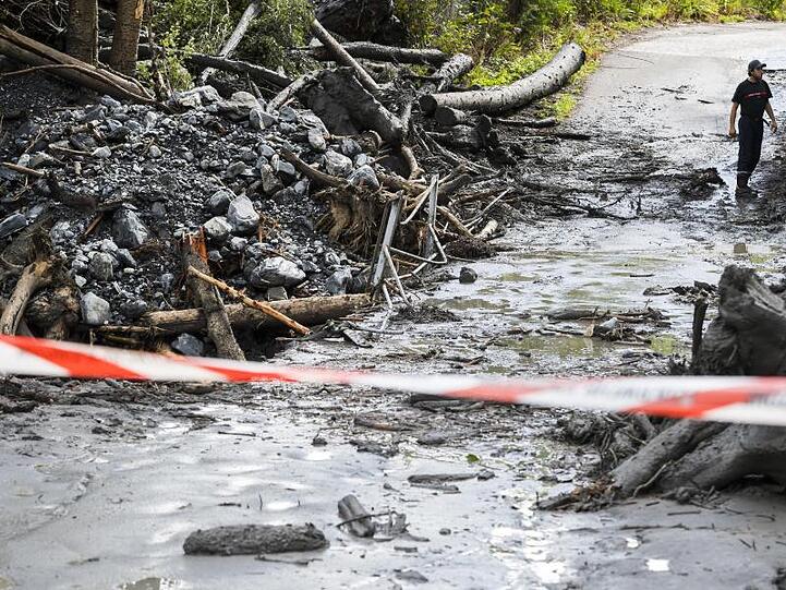 Ein Mann steht auf einer abgesperrten Straße, die von Schlamm, Bäumen und Geröll bedeckt ist. Nach einem heftigen Regen wurde ein Auto durch das Hochwasser des Baches Losentze mitgerissen. In dem Auto befanden sich zwei Menschen, die noch als vermisst gelten.
