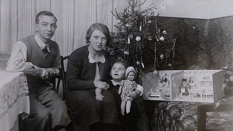 Rosina Schelle 1938 mit ihrer Mutter und ihrem Vater an Weihnachten. Sie hat ein Puppenhaus geschenkt bekommen. Sie sitzen vor dem geschmückten Christbaum, der bereits mit elektrischen Lichtern ausgestattet war. Ihr Vater war technikbegeistert.