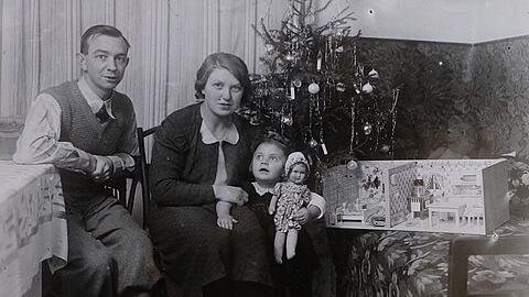 Rosina Schelle 1938 mit ihrer Mutter und ihrem Vater an Weihnachten. Sie hat ein Puppenhaus geschenkt bekommen. Sie sitzen vor dem geschmückten Christbaum, der bereits mit elektrischen Lichtern ausgestattet war. Ihr Vater war technikbegeistert.