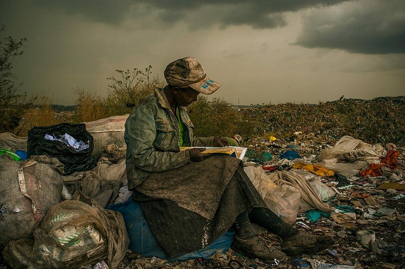Eine Frau sitzt auf der Mülldeponie von Dandora, einem Vorort von Nairobi, Kenia, auf Müll, den sie gesammelt hat. Sie sagt, dass sie beim Müllsammeln als Pause gern mal in Bücher sieht, sogar in Industriekataloge. Diese etwa 8 km vom Zentrum der kenianischen Hauptstadt entfernt liegende Müllhalde, ist eine der größten Afrikas. Die Menschen in den Slums der Umgebung leiden unter einem erhöhten Bleigehalt im Blut und erkranken überdurchschnittlich oft an Krebs- und Nierenkrankheiten. Die aus dem faulenden Müll aufsteigenden Gase führen zu vielen Atemwegserkrankungen. Trotz des hohen Gesundheitsrisikos leben 6.000 bis 10.000 Menschen vom Müll. Sie suchen Lebensmittelreste, Gegenstände, die sie verkaufen können oder Recyclingmaterial. Informelle Kartelle leiten das Recycling und zahlen den Sammlern etwa € 2 am Tag. Die 1975 eröffnete Müllhalde hätte &ndash; internationalen Umweltschutzgesetzen zufolge &ndash; nach 15 Jahren geschlossen werden müssen. 2001 erklärte man, die Halde sei voll, aber sie wird immer noch genutzt.