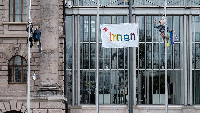 Zwei Menschen haben einen Fahnenmast vor der Bayerischen Staatskanzlei in München erklommen, um gegen das bayerische Genderverbot zu demonstrieren.