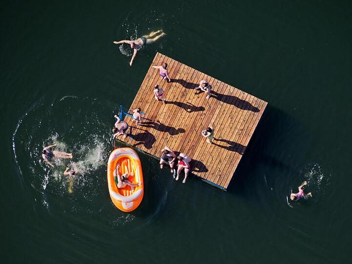 Kinder toben auf einer Holzinsel und in einem Schlauchboot in einem See (Luftaufnahme). Foto: Julian Stratenschulte/Archivbild