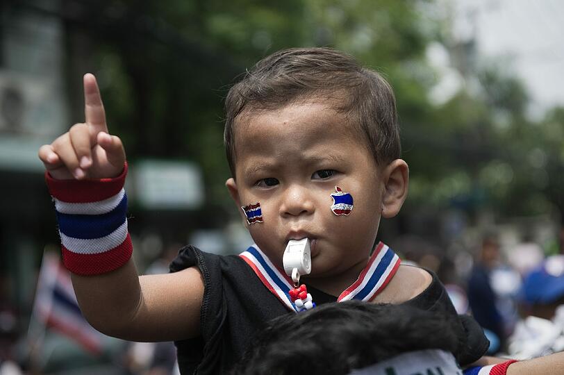 Ein kleiner Junge mit Schminke in Landesfarben und Pfeife im Mund sitzt auf den Schultern eines Demonstranten in Bangkok. In Thailand gehen die Proteste gegen die Regierung weiter, nachdem am 1. April ein Demonstrant erschossen wurde.