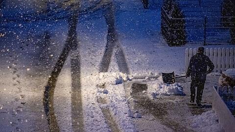 Ein Mann geht am frühen Morgen mit einer Schneeschaufel auf einer zugeschneiten Straße.