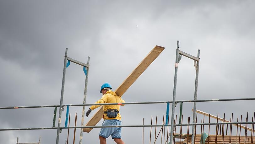 Ein Bauarbeiter trägt auf einer Baustelle auf einem Hausdach ein Brett.