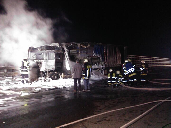 Im Hintergrund beleuchten die Scheinwerfer der Feuerwehr gespenstisch den Qualm, der aus dem ausgebrannten Lkw steigt.