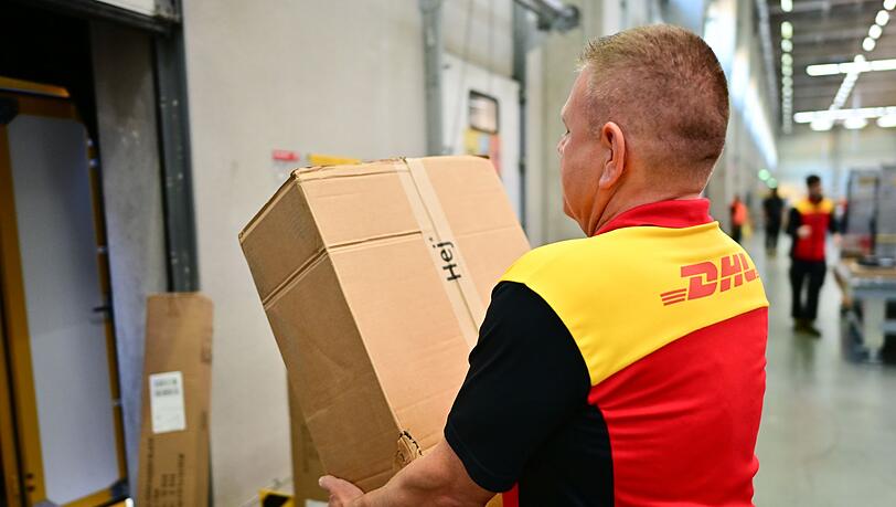 Ein Paketbote schleppt in einem DHL-Standort ein schweres Paket. (Archivbild)