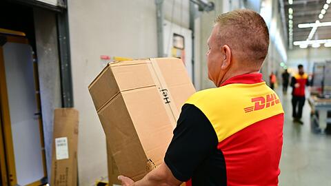 Ein Paketbote schleppt in einem DHL-Standort ein schweres Paket. (Archivbild)